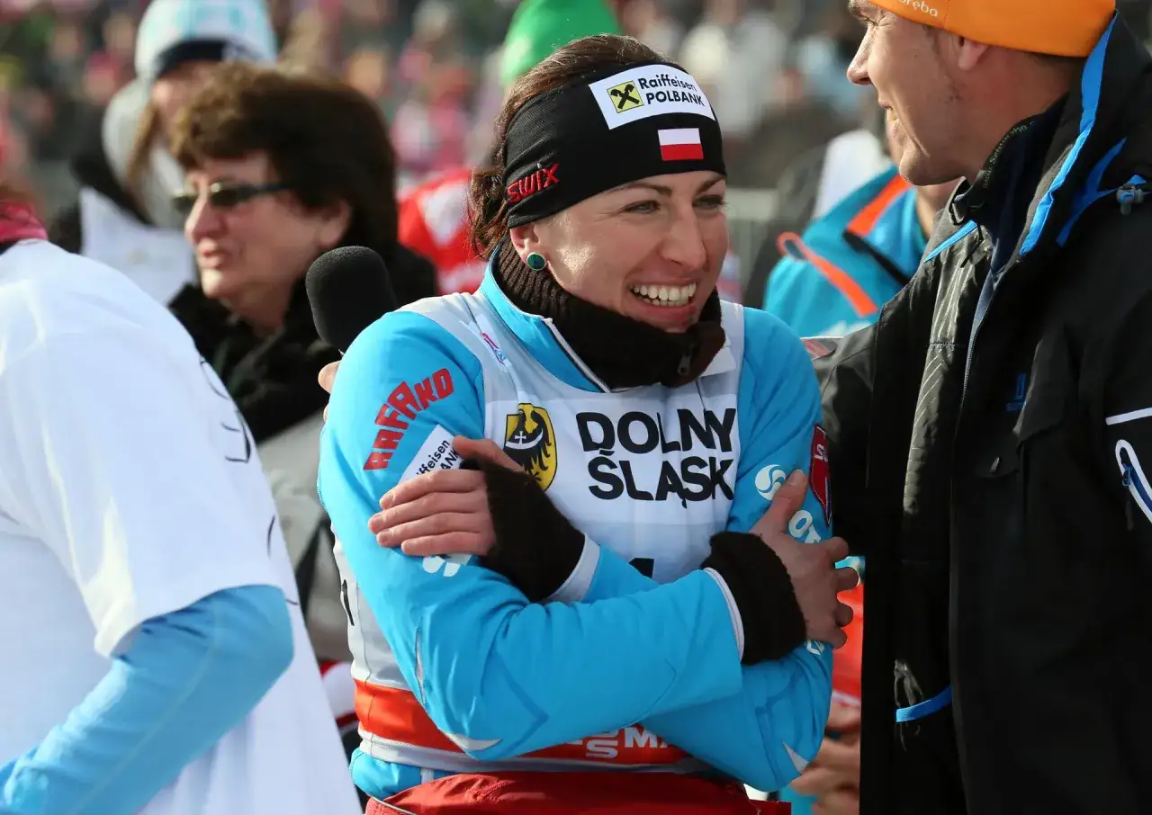 Justyna Kowalczyk i Sebastian Parfjanowicz podczas zawodów. Justyna uśmiecha się, ubrana w niebieską kurtkę z logo