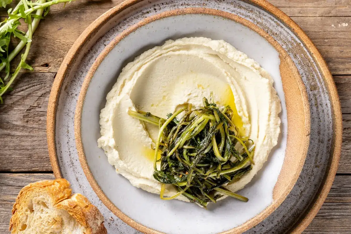 Cremoso pur&egrave; di fave, condito con olio e verdure verdi saltate, servito su un piatto rustico con pane croccante.