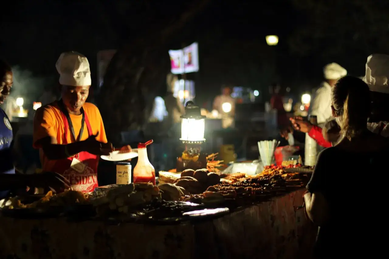 Nocny targ na Zanzibarze. Sprzedawca w czapce kucharskiej podaje talerz z pysznym zanzibarskim jedzeniem.