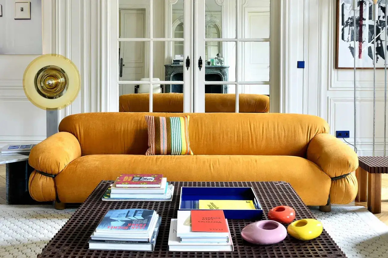 Soggiorno con divano giallo ocra, cuscino a righe colorate e tavolino con libri e oggetti decorativi. Abbinamenti audaci e moderni.