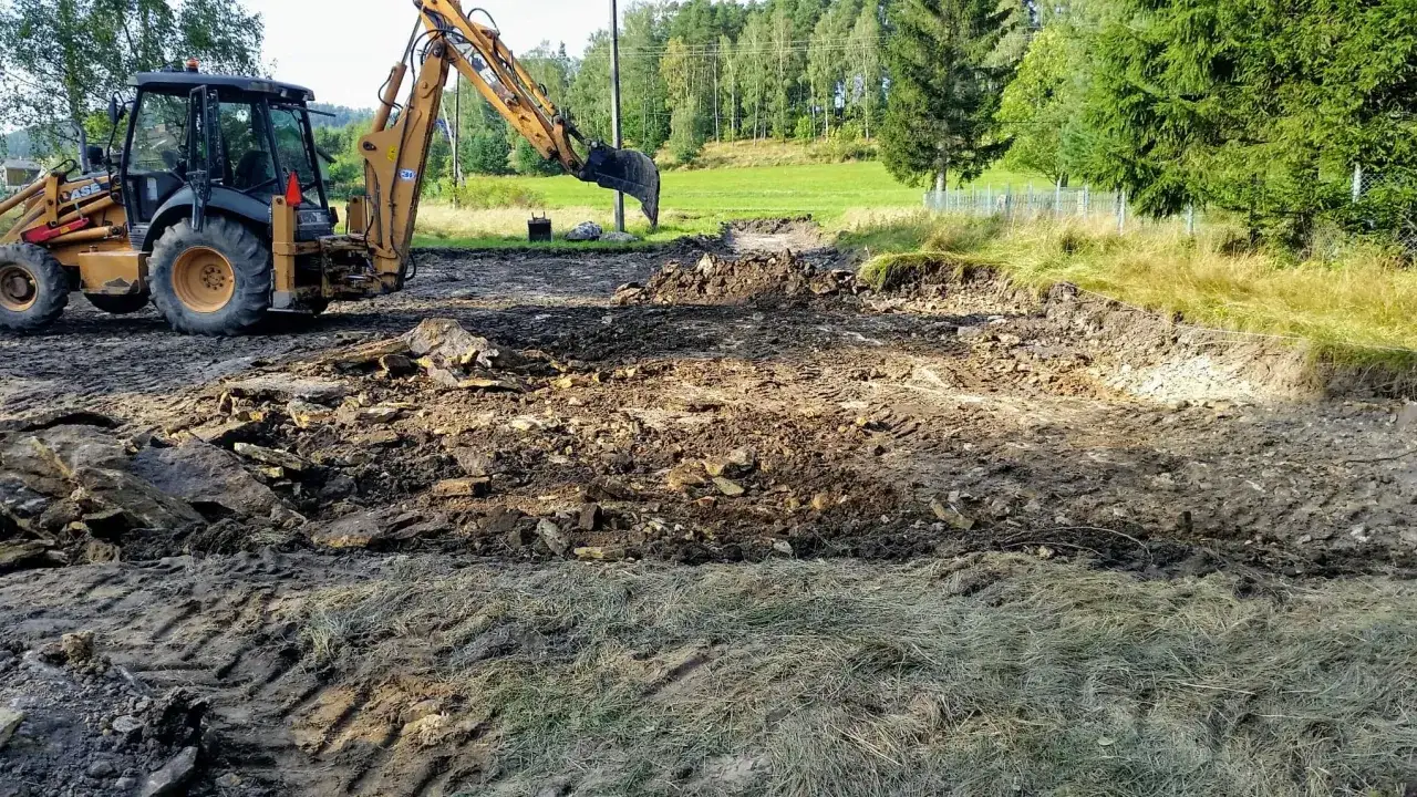 Koparka pracuje przy wykopie pod fundament. Teren jest błotnisty, z gruzem i kamieniami.