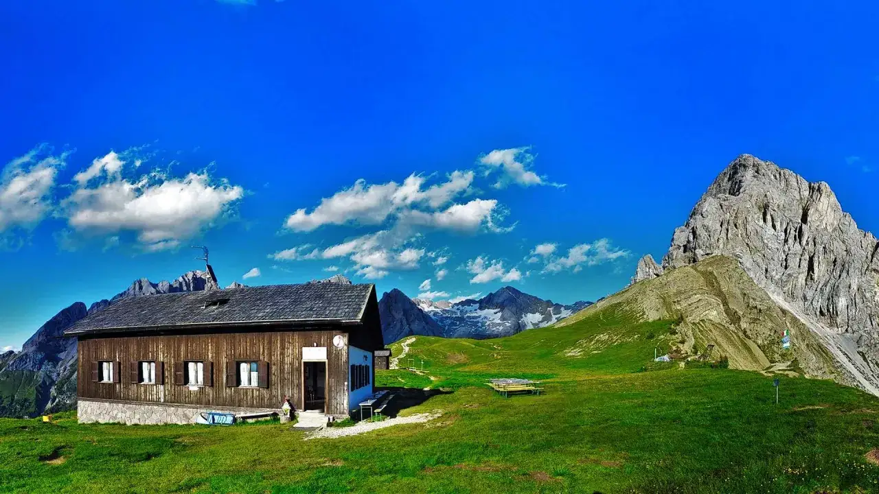 Rifugio Passo San Nicolò, un'accogliente baita in legno immersa nel verde, con imponenti vette sullo sfondo e un cielo azzurro punteggiato di nuvole.