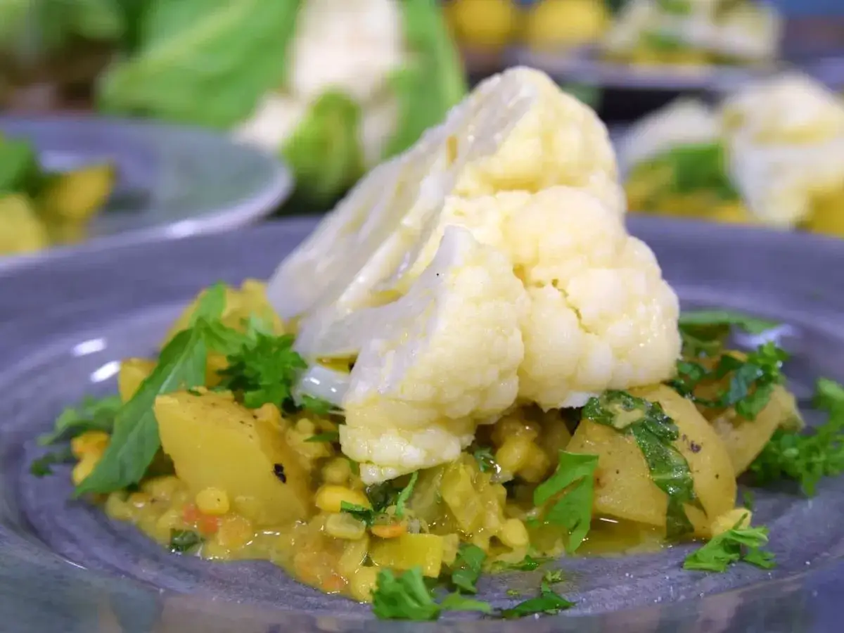 En n&auml;rbild p&aring; en tallrik med en gul currygryta med potatis och majs, toppad med &aring;ngad blomk&aring;l och f&auml;rska &ouml;rter. Perfekt f&ouml;r att koka blomk&aring;l till en smakrik m&aring;ltid.