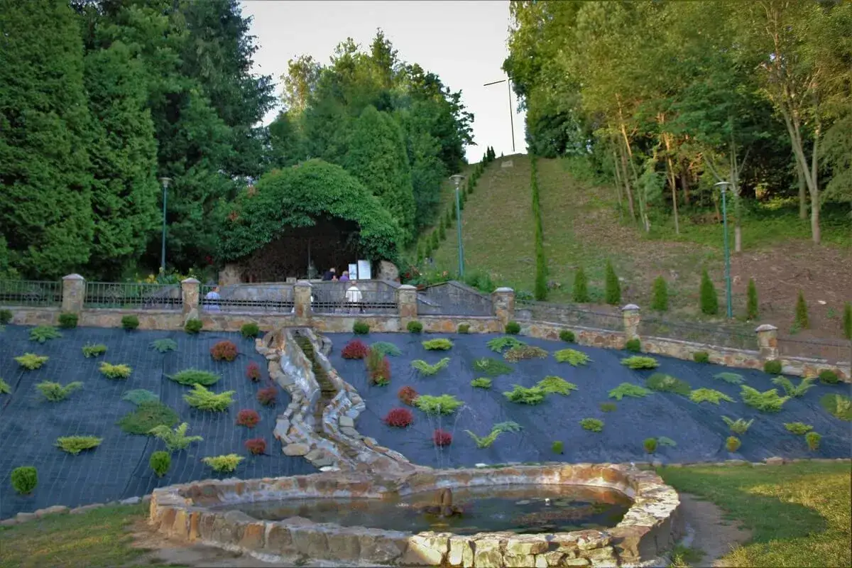 Cudowne źródełko w Zwierzyniu, trasa pielgrzymkowa. Woda spływa kaskadami po kamieniach, otoczona zielenią i ozdobnymi krzewami.