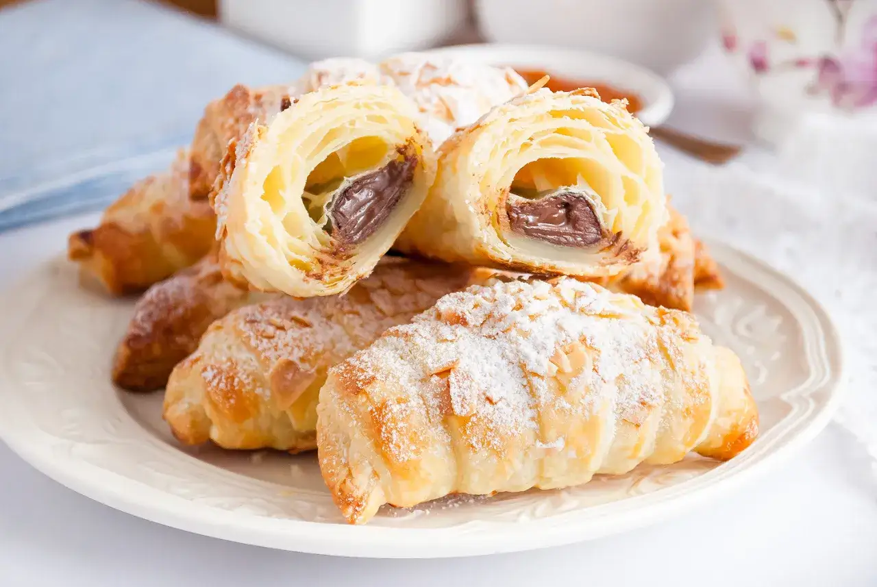 Chrupiące croissanty z ciasta francuskiego z nadzieniem czekoladowym, posypane cukrem pudrem i płatkami migdałów. Idealne na słodkie śniadanie.