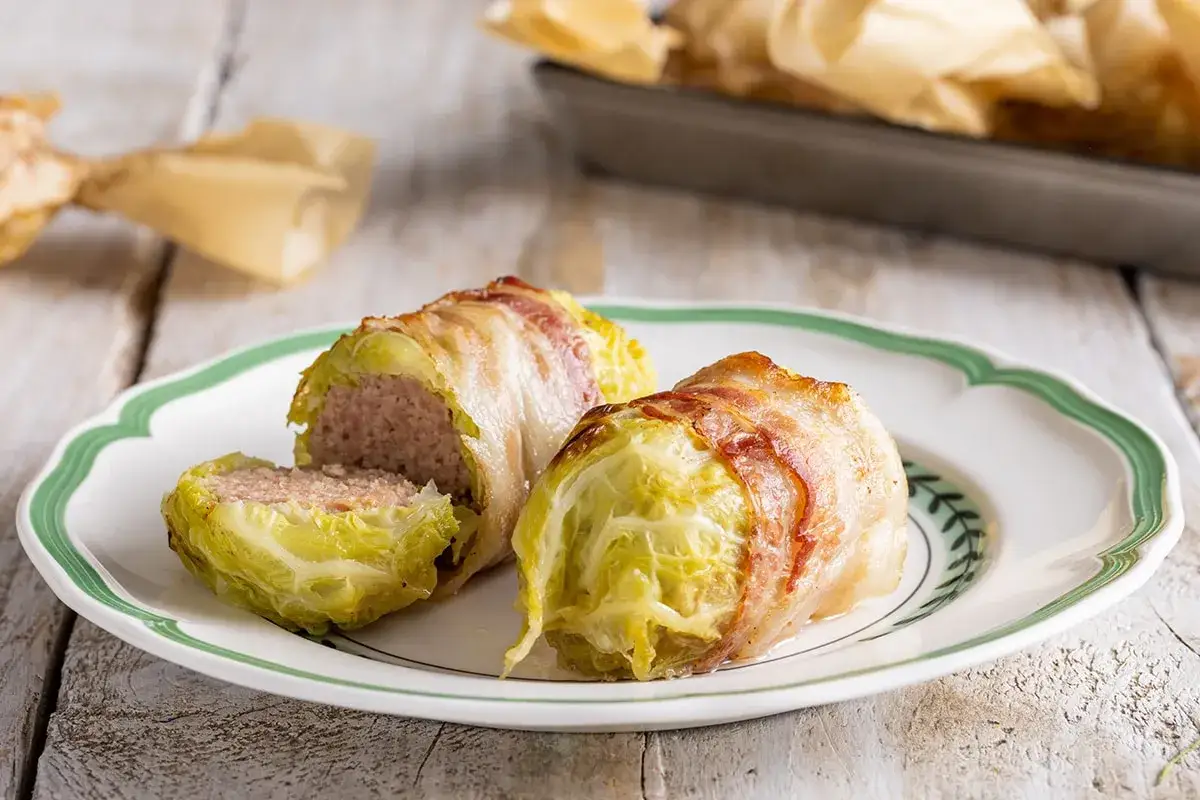 Involtini di verza con carne, avvolti in pancetta croccante, su un piatto bianco con bordo verde. Uno &egrave; tagliato a met&agrave;, mostrando il ripieno succulento.