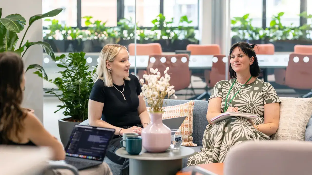 Three women in a modern office setting, discussing ideas. Their engaged expressions suggest positive employee sentiment.