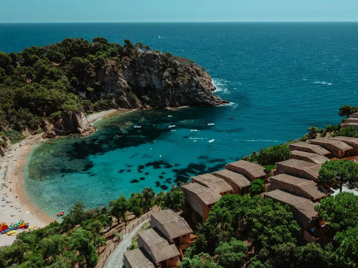 Vista a&eacute;rea de una cala turquesa con casas de **Pola Giverola Resort** y pinos verdes.
