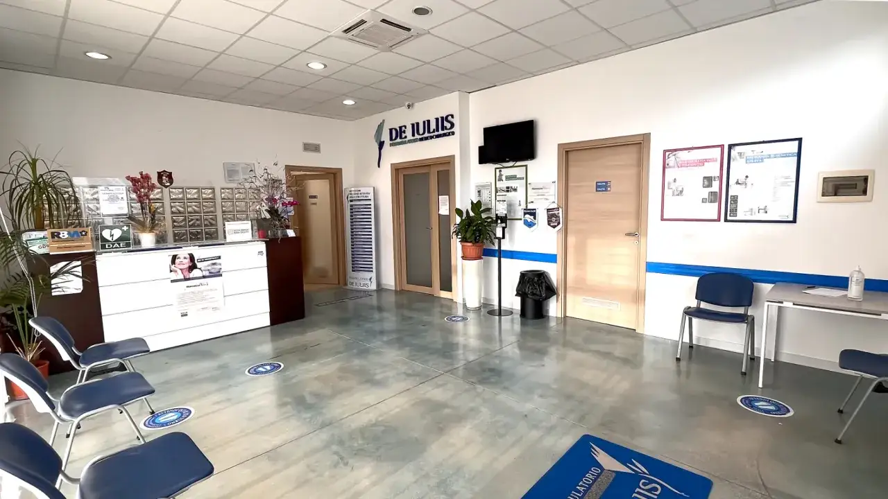 Sala d'attesa di un poliambulatorio con bancone reception, sedie blu e cartellonistica. La segretaria poliambulatorio accoglie i pazienti.