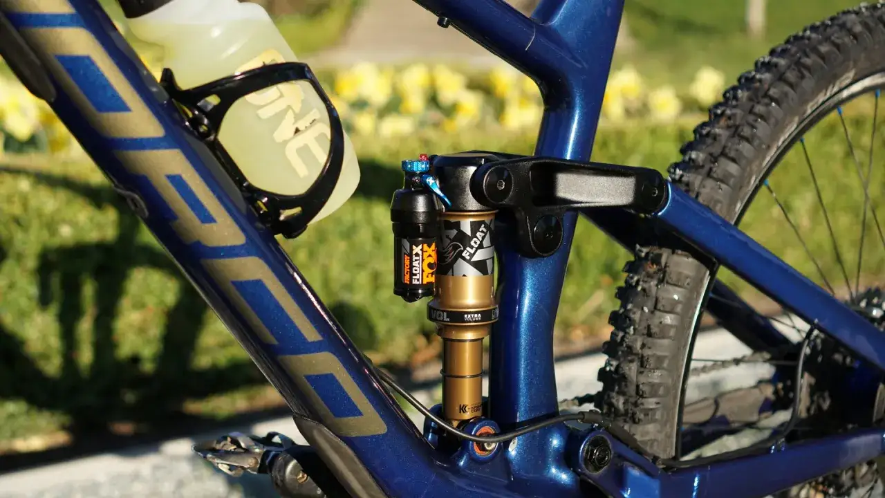 Close-up of a blue mountain bike frame, featuring a Fox Float X Factory shock absorber. The bike has a water bottle cage with a yellow bottle.