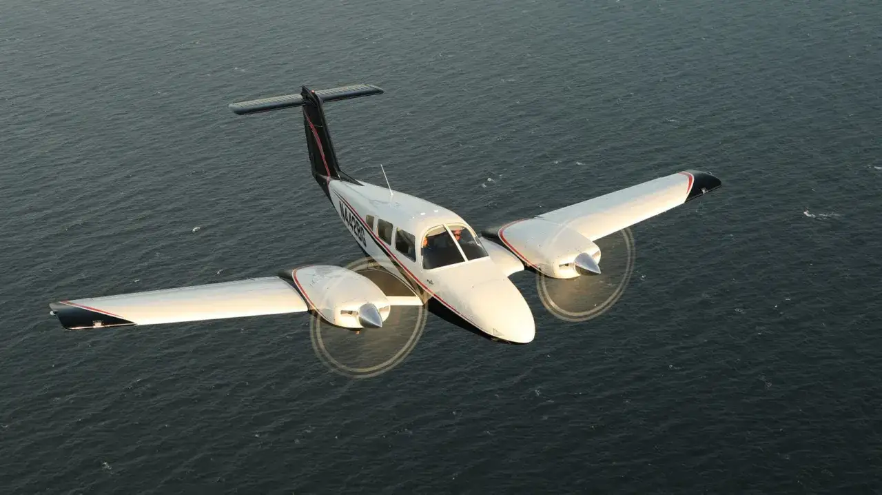 Eine Piper PA-23 Aztec, ein zweimotoriges Flugzeug, fliegt über ruhigem Wasser.