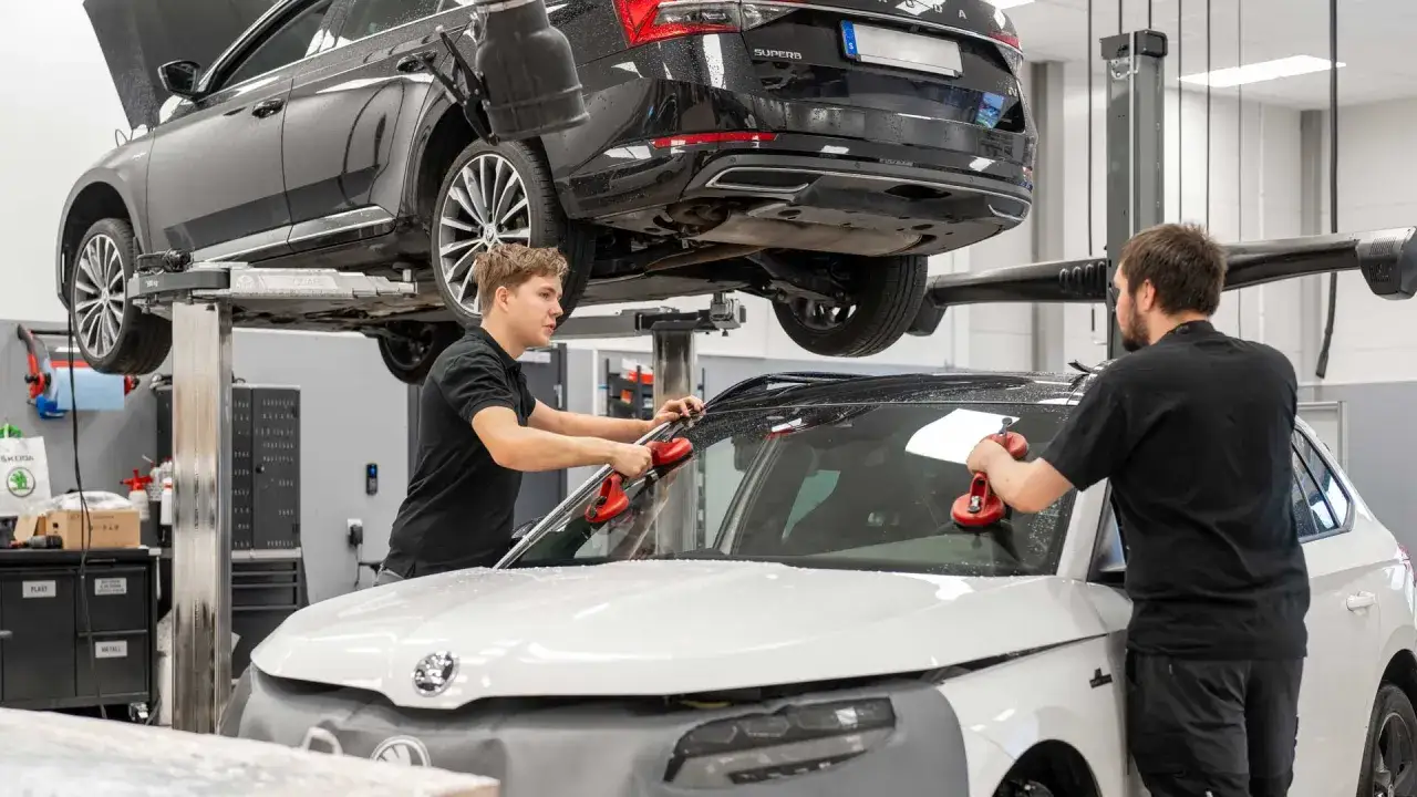 Två mekaniker arbetar med en Skoda Octavia på en verkstad. En bil lyfts upp på en lyft, medan en annan bil har vindrutan borttagen. Detta är en del av serviceintervallet för Skoda Octavia.