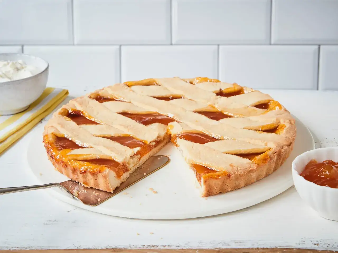 Ein k&ouml;stliches Crostata Rezept mit Aprikosenmarmelade, das auf einem wei&szlig;en Tisch serviert wird. Daneben eine Sch&uuml;ssel mit Marmelade und eine mit Sahne.