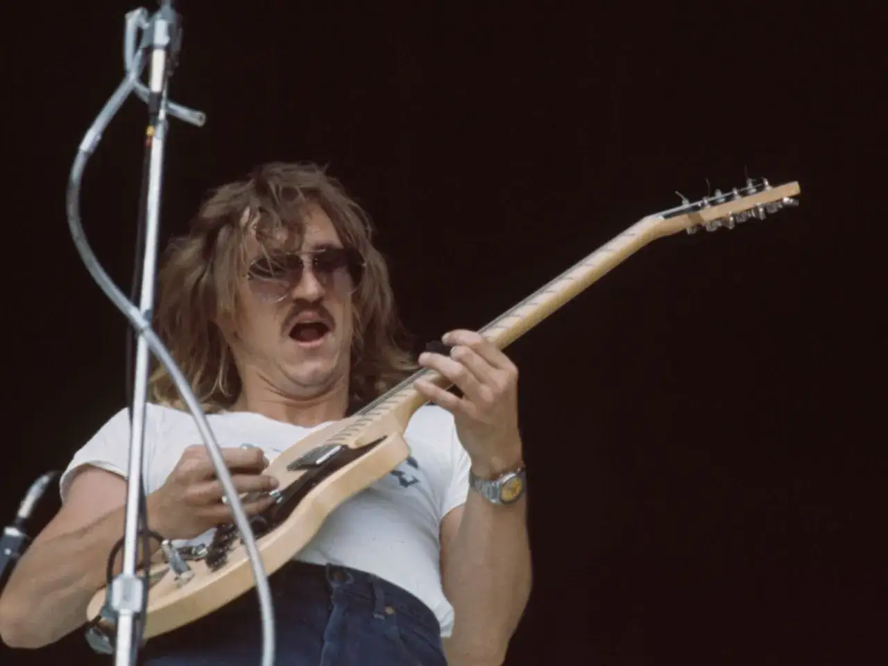 Muzyk z wąsem i okularami przeciwsłonecznymi gra na gitarze. Jego usta są otwarte, jakby krzyczał do mikrofonu, tworząc efektowny **talk box**.