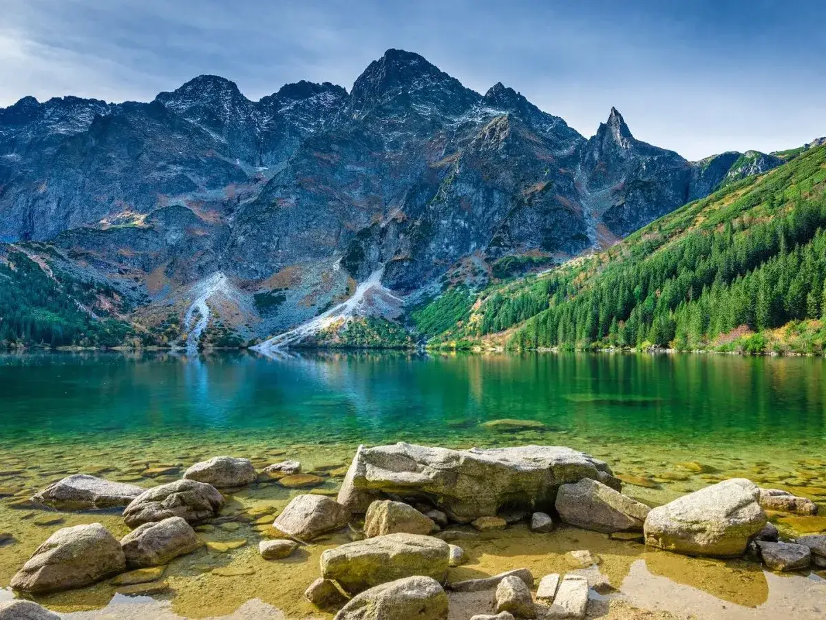 Bergsee mit klarem Wasser und Felsen im Vordergrund, umgeben von gr&uuml;nen W&auml;ldern und schroffen Gipfeln. Ein wundersch&ouml;ner Anblick in Polen.
