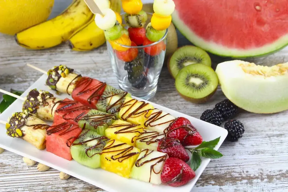 Brochetas de frutas frescas con chocolate y pistachos, acompañadas de sandía, melón, kiwi y plátano.