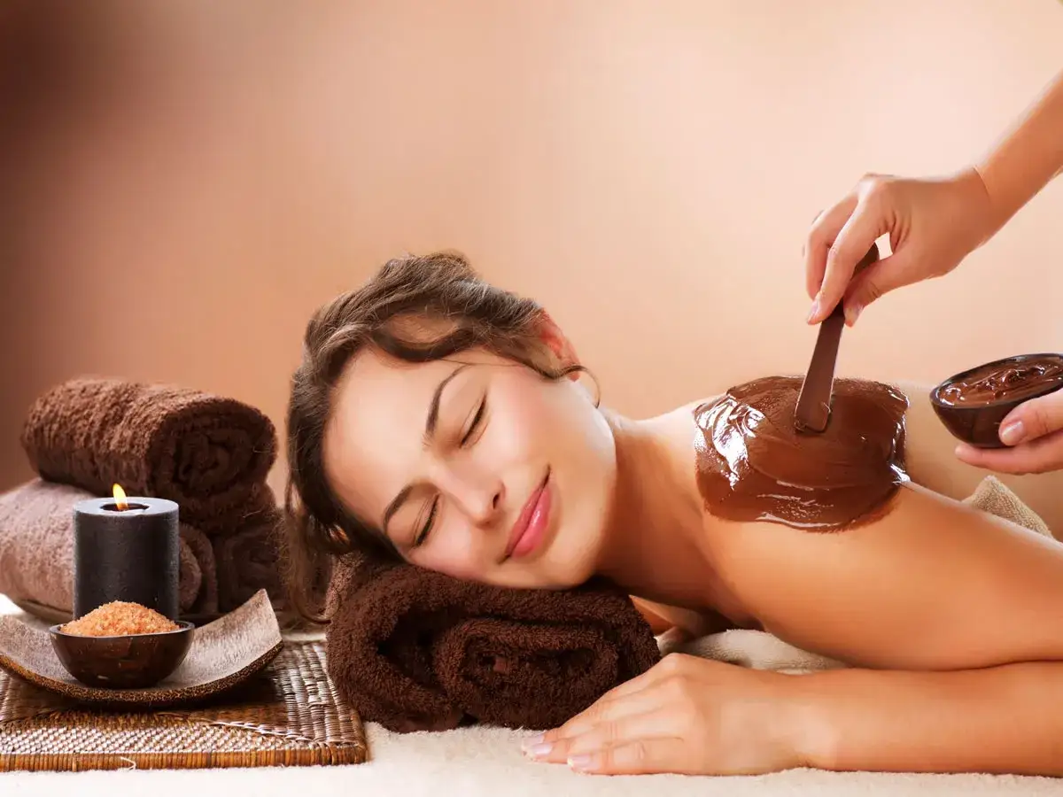 Una mujer disfruta de un relajante ba&ntilde;o de chocolate spa mientras le aplican una mascarilla de cacao en la espalda.
