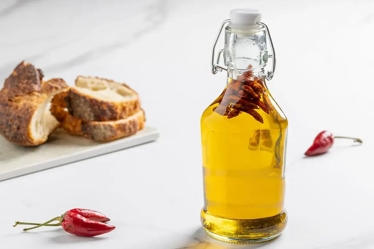 Olio al peperoncino in bottiglia, con pane affettato e peperoncini rossi sullo sfondo.