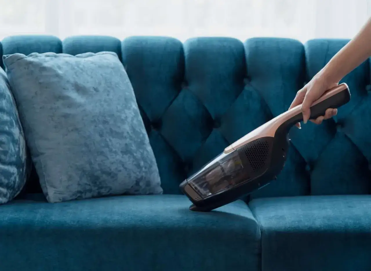 A hand uses a cordless vacuum to clean a plush teal velvet sofa, showing how to wash velvet furniture gently.