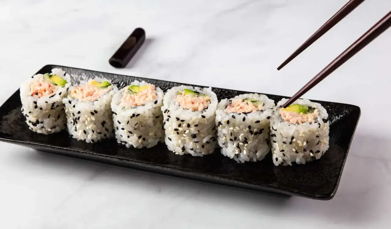 Siedem kawałków california sushi z awokado i łososiem, posypane sezamem, na czarnym talerzu. Pałeczki sięgają po jeden z nich.