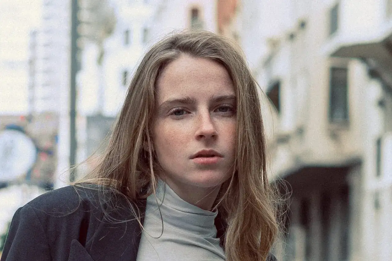 Jeune femme au regard intense, ses cheveux ch&acirc;tain clair cendr&eacute; ou glac&eacute; encadrent un visage parsem&eacute; de taches de rousseur. Elle porte un col roul&eacute; gris et une veste sombre.