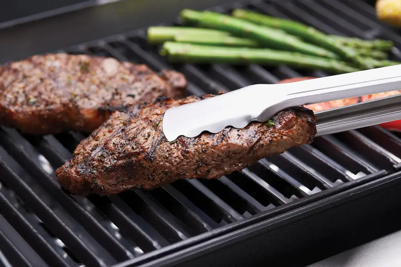 Des steaks juteux et des asperges grillent sur la grille BBQ. Des pinces retournent un steak parfaitement cuit.