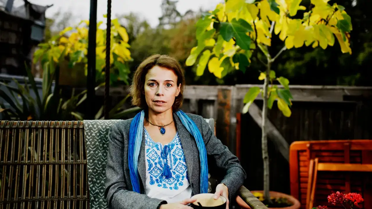 Anna Borgeryd sitter i en korgstol utomhus, med en bl&aring; scarf och en broderad blus. En kaffekopp vilar i hennes h&auml;nder.
