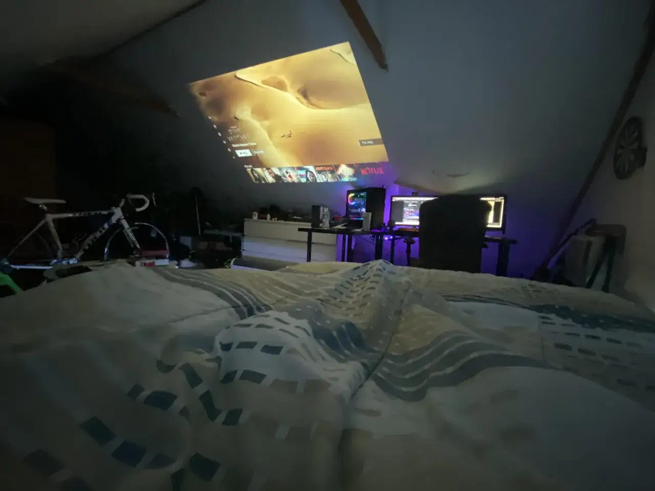 Przytulny pok&oacute;j gamingowy na poddaszu z projektorem wyświetlającym Netflix. Obok ł&oacute;żka stoi rower.