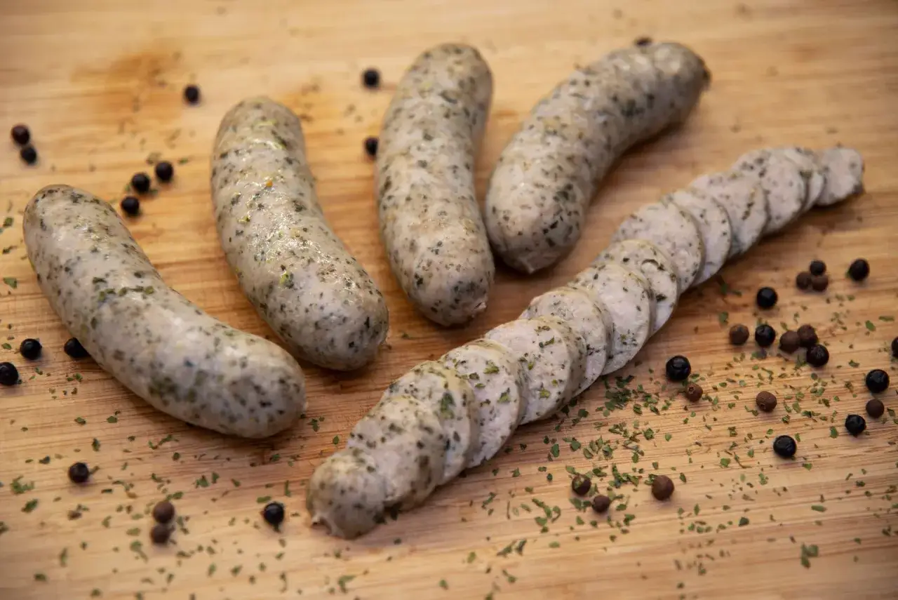 Gotowy przepis na białą kiełbasę. Kilka sztuk ułożonych na desce, jedna pokrojona w plastry, z dodatkiem pieprzu i ziół.
