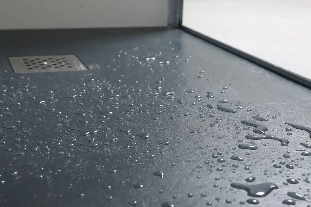 Gotas de agua sobre plato de ducha de resina. Un desag&uuml;e met&aacute;lico se ve en la esquina superior izquierda.