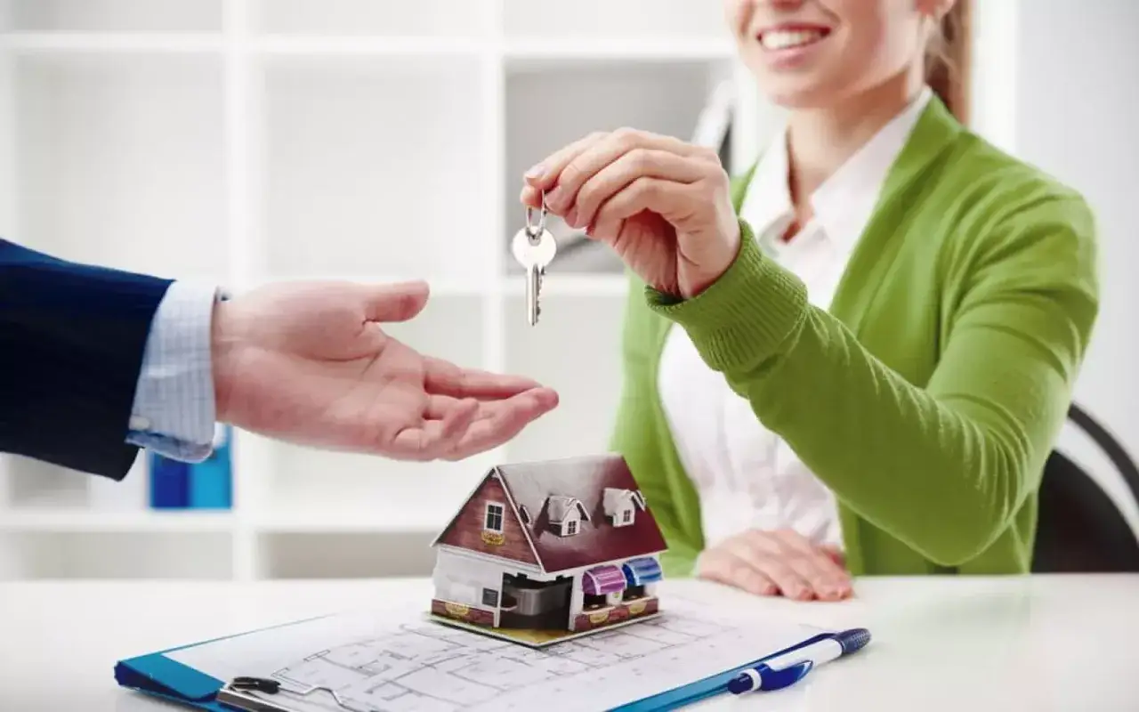 Une main tend une clé pour un transfert ptz, symbolisant l'acquisition d'une maison. Une maquette de maison et des plans sont sur la table.
