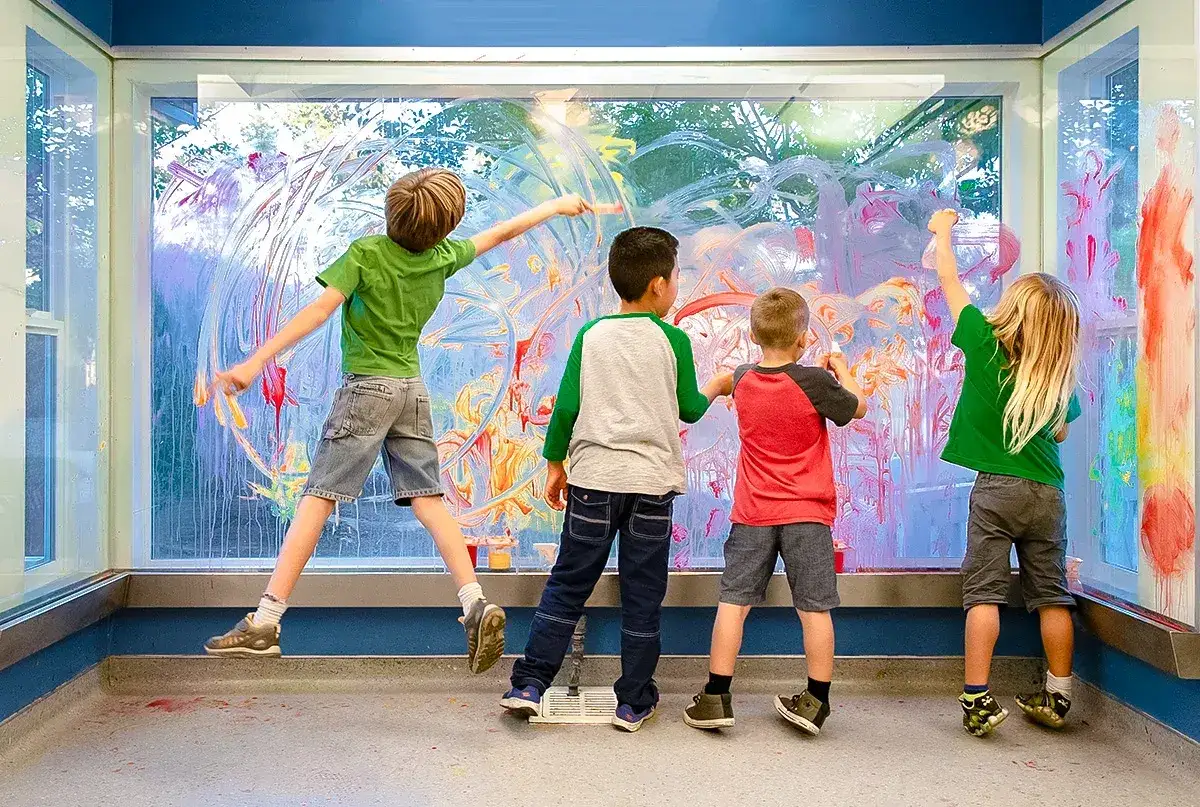 Niños creando arte en infantil pintando un gran ventanal con colores vibrantes.