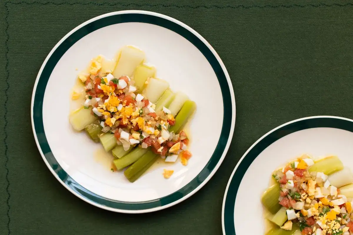 Puerros a la vinagreta servidos en platos blancos con borde verde, cubiertos con una mezcla de tomate picado, huevo duro y perejil.