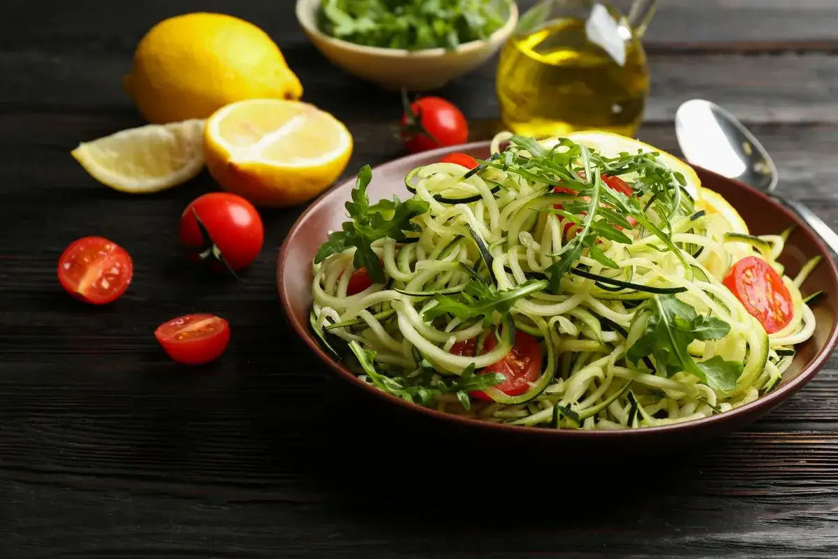 Un piatto di spaghetti di zucchine freschi, guarniti con pomodorini e rucola, pronti per essere conditi con olio d'oliva e limone.