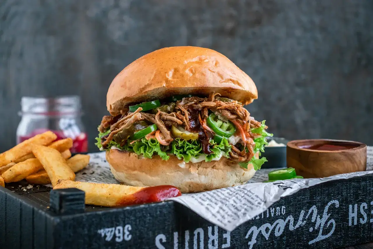 Saftiger pulled pork burger mit Salat, Jalapeños und Gurken. Die Zutaten für diesen Burger sind perfekt aufeinander abgestimmt. Dazu Pommes und Dip.