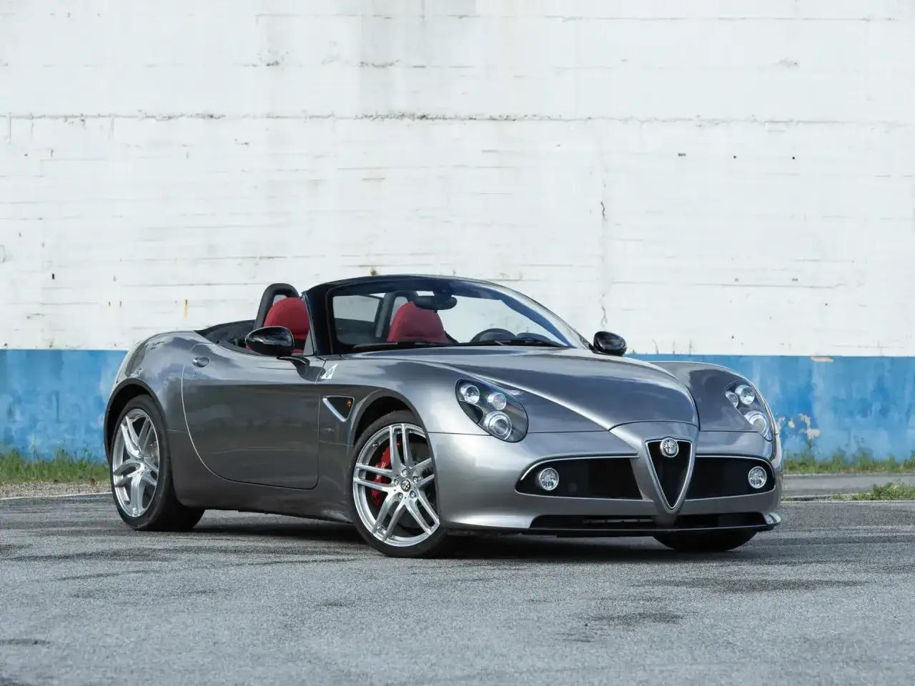Szary kabriolet Alfa Romeo 8C z czerwonymi siedzeniami stoi na tle białej ściany.