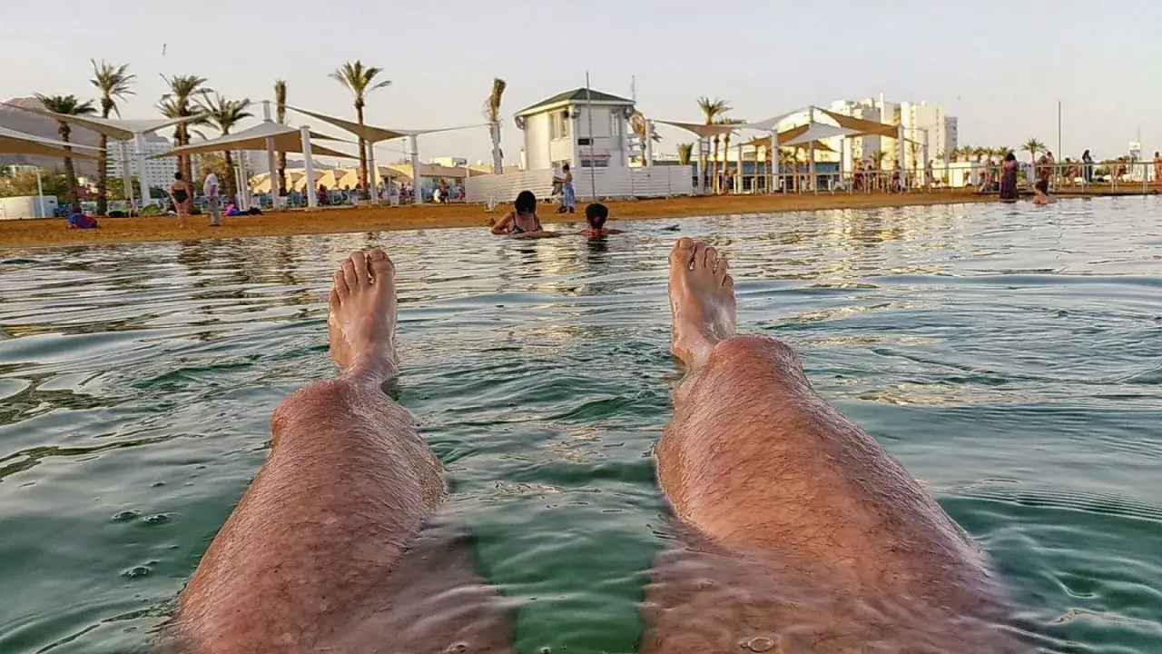 Nogi unoszą się na Morzu Martwym podczas wycieczki do Izraela. W tle plaża, palmy i budynki.