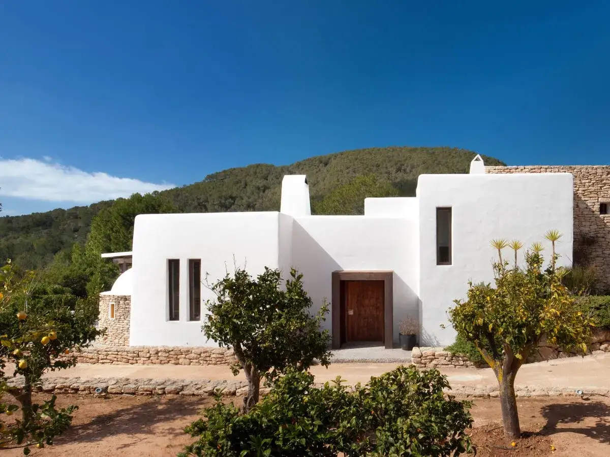 Exterior de una casa ibicenca blanca, con puerta de madera y rodeada de árboles frutales bajo un cielo azul.