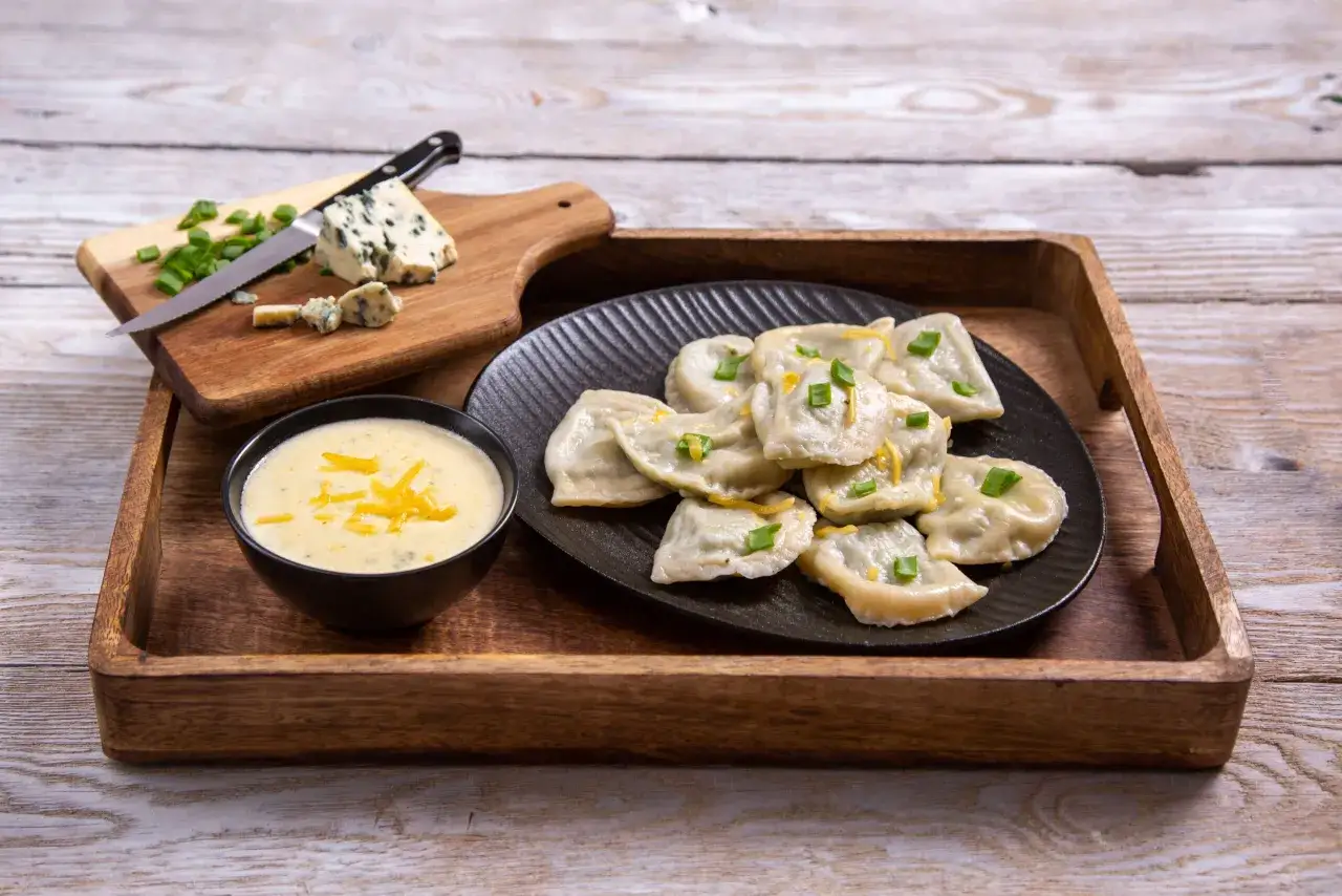 Pyszne pierogi ze szpinakiem i serem feta, podane z kremowym sosem. Obok deska z serem pleśniowym i szczypiorkiem.