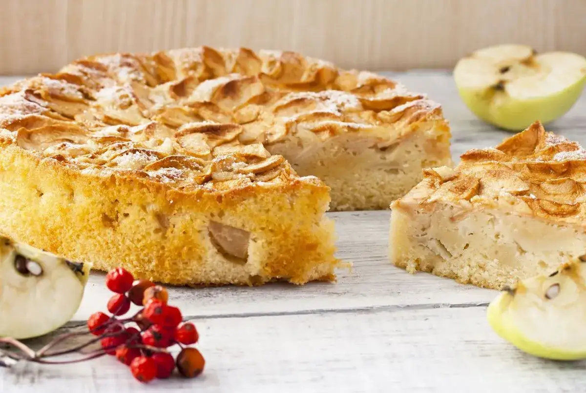 Un g&acirc;teau compote de pomme sans sucre, saupoudr&eacute; de sucre glace, avec des tranches de pomme sur le dessus. Des morceaux de pomme et des baies rouges d&eacute;corent la table.