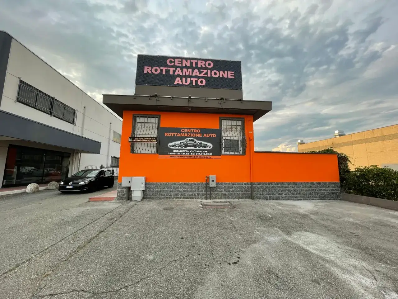 Centro rottamazione auto con insegna luminosa e un'auto nera parcheggiata. Servizi di autodemolizione.