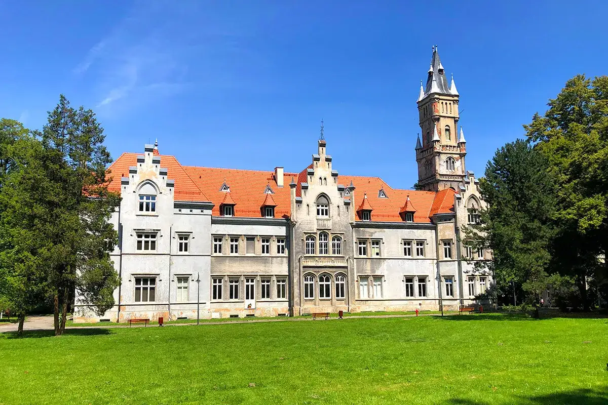Imponujący pałac w Nakle Śląskim z charakterystyczną wieżą i pomarańczowym dachem, otoczony zielonym trawnikiem i drzewami.