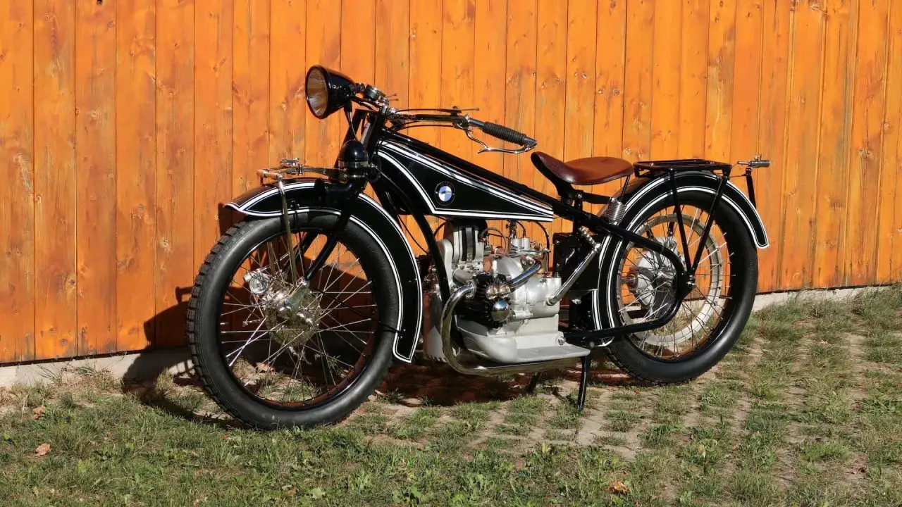 Klasyczny motocykl BMW R32, ikona BMW historii, z charakterystycznym silnikiem typu bokser i skórzanym siodłem, na tle drewnianego płotu.