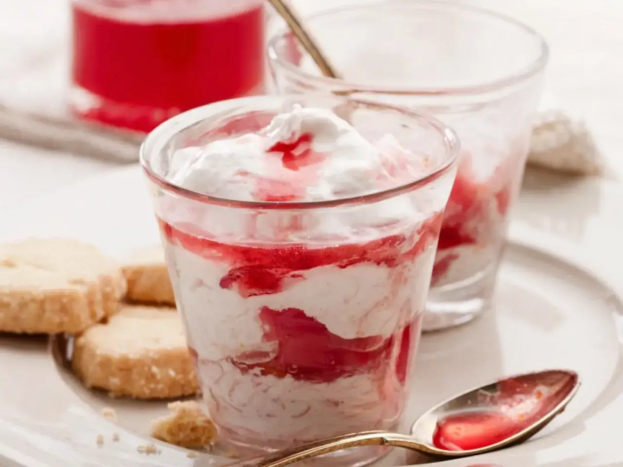 Cremiges Dessert mit Rhabarberkompott und Sahne, serviert mit Keksen. Ein L&ouml;ffel mit roter Sauce liegt daneben.