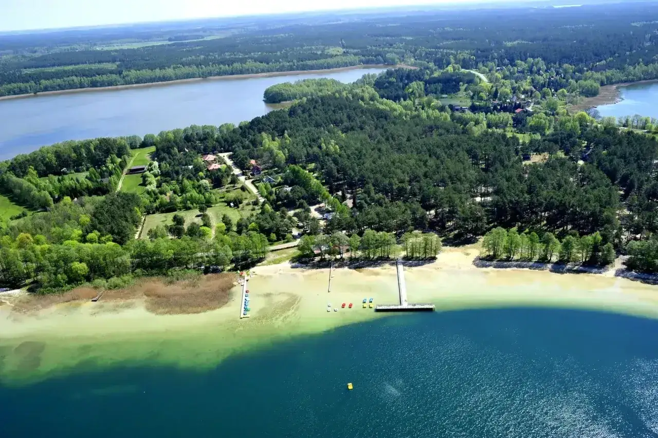 Malownicze jezioro Warchały otoczone lasem, z piaszczystą plażą, pomostami i kolorowymi łódkami.