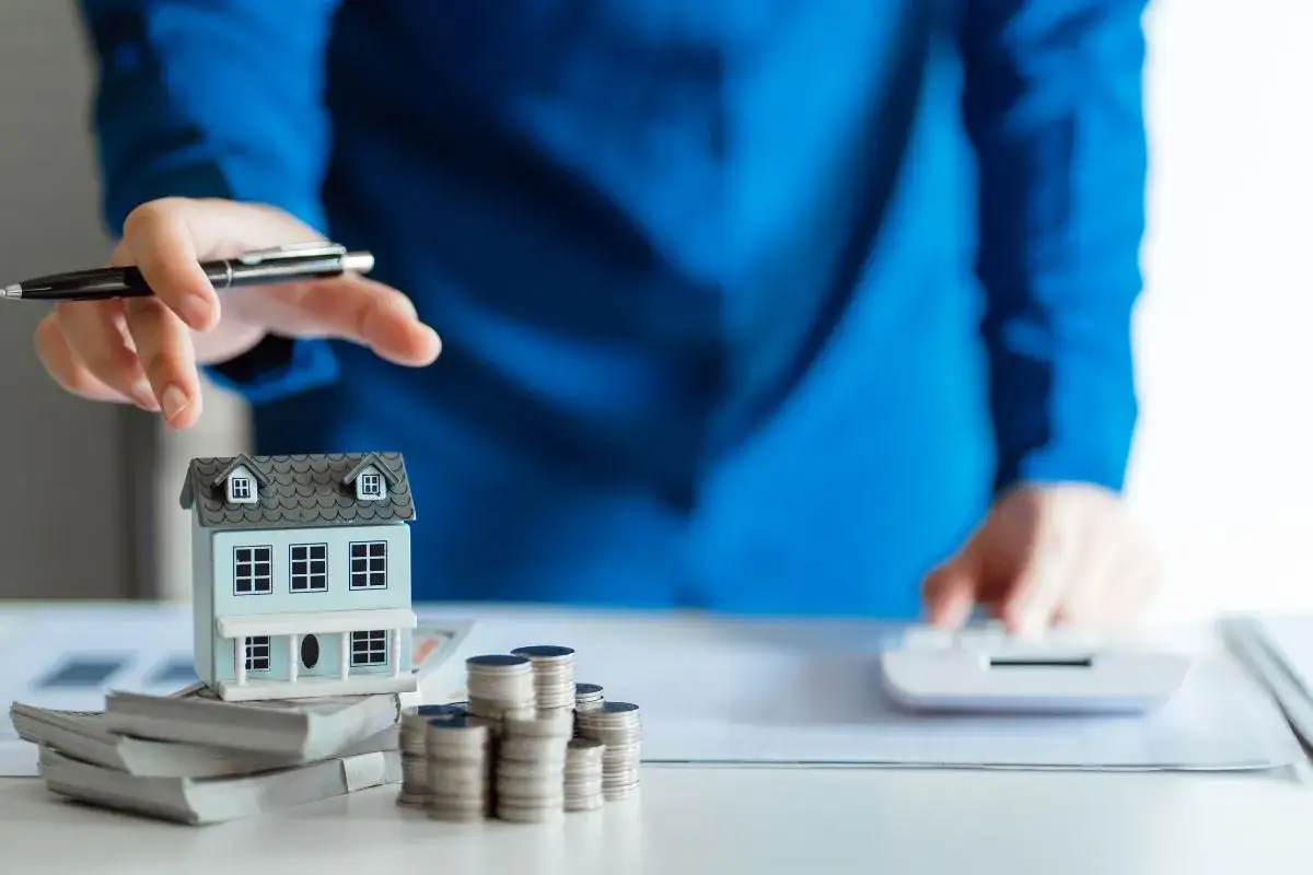 Miniatura di una casa su pile di denaro e monete, con una mano che tiene una penna. Simboleggia l'acquisto con permuta e la pianificazione finanziaria.