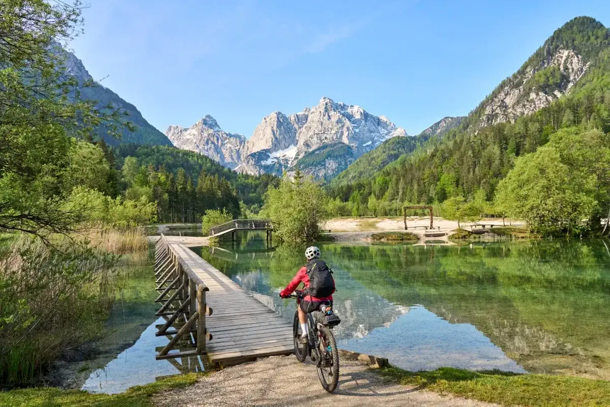 A cyclist enjoys a scenic ride on a wooden bridge over a clear lake, with majestic mountains in the background. Perfect for cycling tours Slovenia adventures.