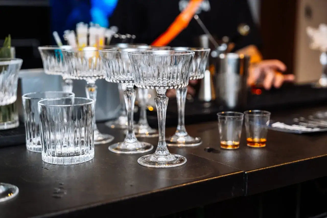 Eleganckie kieliszki i szklanki czekają na gości. W tle widać rękę przygotowującą drinki, jakby to był drink bar zamiast barmana.