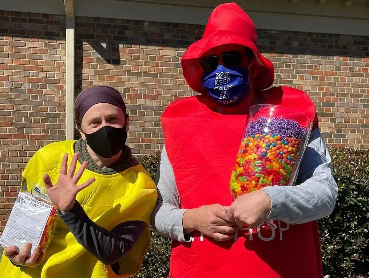 Two people in costumes celebrate Purim meaning with candy. One holds a jar of colorful jelly beans, the other a package of hamantaschen.