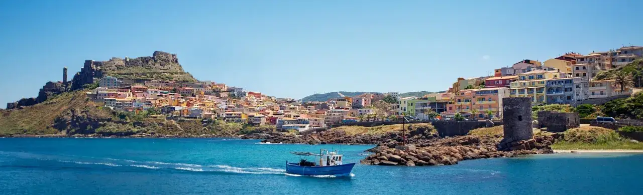 Vue panoramique d'une ville côtière colorée en Sardaigne, avec un bateau bleu naviguant sur la mer turquoise. Idéal pour savoir que faire en Sardaigne.